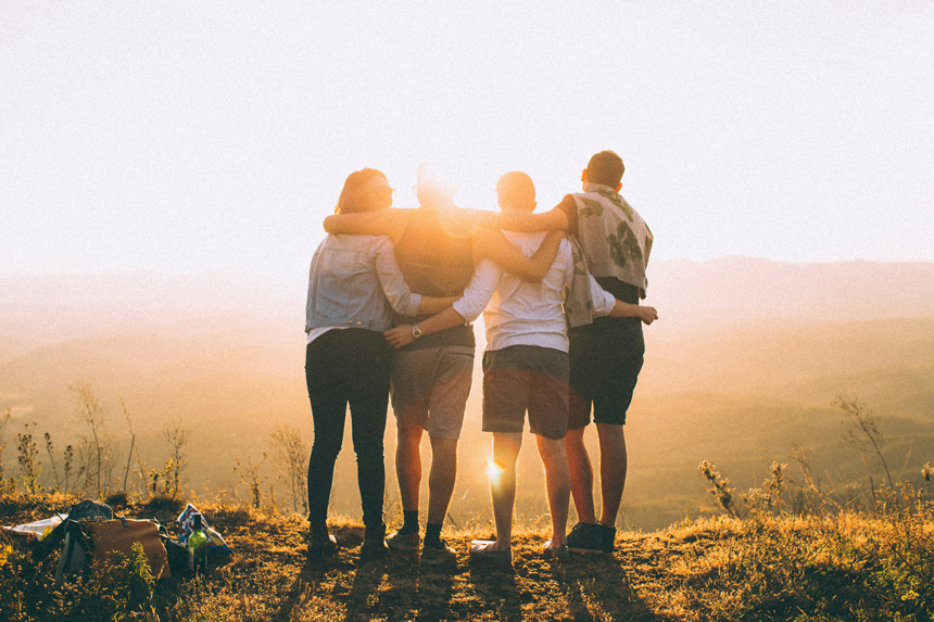 A photo of the backs of a young family of four with their arms stretched across each other backs staring off into the golden hour sun on a cliff.