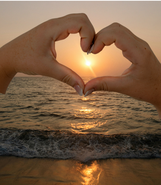 Two hands creating a heart to bring focus to the bright setting sunset at the beach.
