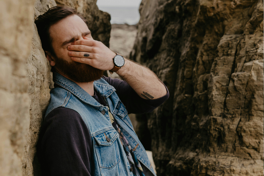 A photo of a young man pinching his eyes while leaning on a tall rock settlement. He is overwhelmed with the weight of the things he has witnessed.