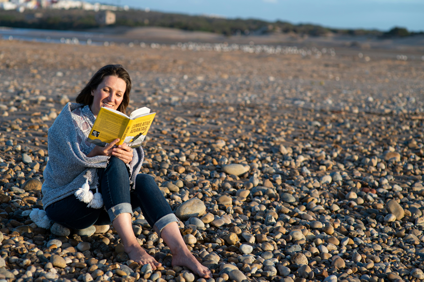A photo of a women reading a yellow book at the beach, that features a rocky shore like Lake Superior. She is smiling with contentment as she embraces the new knowledge.
